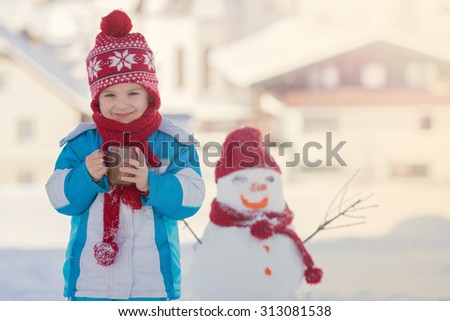 Happy beautiful child building snowman in garden, winter time, holding cup of hot tea, smiling at camera