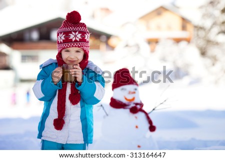 Happy beautiful child building snowman in garden, winter time, holding cup of hot tea, smiling at camera