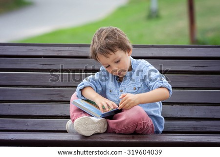 Cute boy, read a book in the park, sitting on bench, summertime. Education for children