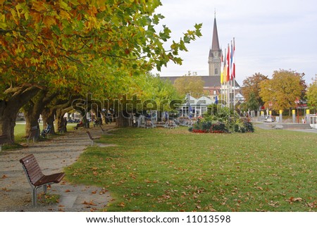 People rest on benches under plane trees in autumn foliage in a park in Radolfzell, Germany. Across the lawn is a cafe, framed by colorful flags and a church tower in the background.