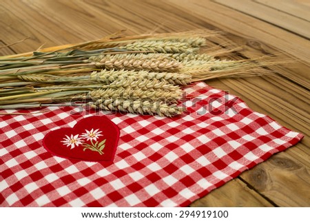 Some wheat, rye, barley ripe cereal ears on a background of red and white checkered tablecloth with a red heart and two embroidered white Edelweiss on rustic wooden table surface