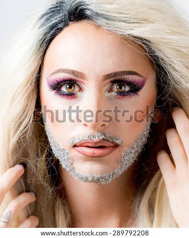 Glamorous man in drag queen make-up with heavy mascara on staring eyes, glittery beard, wild hair on white background.