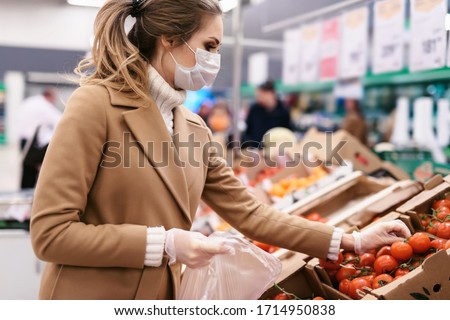 Shopping during the coronavirus Covid-19 pandemic. A young woman buys tomatoes in a supermarket. Woman in facial mask and gloves to prevent infection.