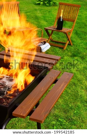 Picnic Scene. Flaming BBQ Grill, garden furniture, basket and wine.