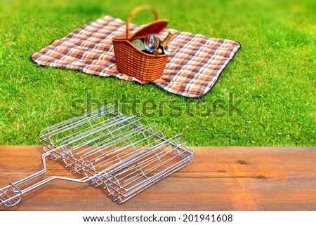 Summer Picnic on Lawn Scene. BBQ Grid on wood table. Basket with wine and wineglass on the blanket in blurred background.