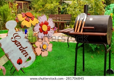 Weekend Summer Backyard Party Or Picnic Concept. Sign Garden Close-up. BBQ Grill With Tools, Outdoor Wood Furniture Plants And House On The Blurred Background