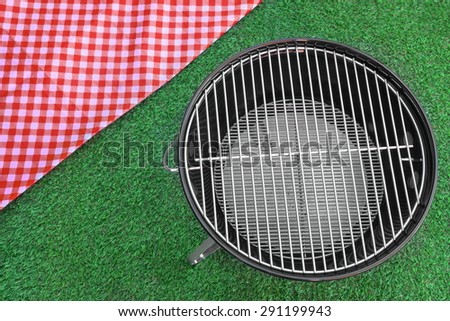 Overhead View Of Red Picnic Tablecloth And Empty BBQ Grill Appliance On The Summer Green Lawn Background
