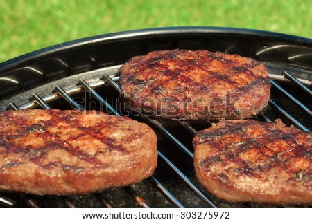 Close-up Of BBQ Hamburger Patties On The Hot Portable Charcoal Grill. Backyard Grass In The Background. Cookout Food For Summer Weekend Picnic Or Party.