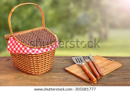 Close-up Of Picnic Bench With Hamper And BBQ Grill Tools And Summer Garden With Sunbeams In The Background
