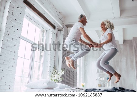 Photo of Love lives forever! Senior couple at home. Handsome old man and attractive old woman are enjoying spending time together. Having fun and jumping in bed.