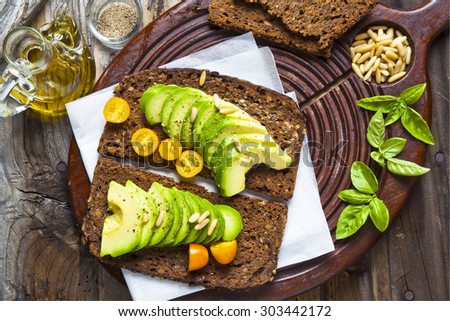 sandwich with rye bread on a board for cutting: avocado, yellow tomatoes, pine nuts and basil leaves. grated black pepper and olive oil. on the old wooden background.