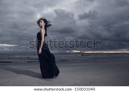 Beautiful brunette model in long black dress standing on the beach. Cloudy weather. Sunset