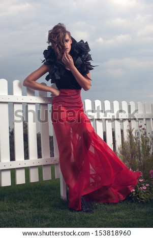 fashionable model posing near white fence. Long red and black dress. Red skirt.  black body. volume accessory on his shoulders. black shoes with spikes