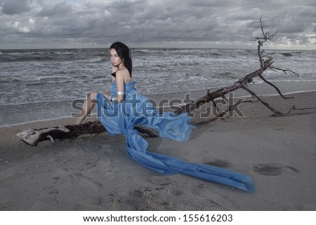 Beautiful brunette model sitting on old tree branch on the beach during storm. Vogue style. Long blue skirt