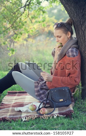 Hipster style young woman using tablet pc and eating an apple during a rest in the autumn park