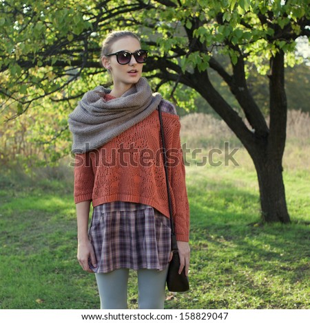 Trendy model in knitted sweater, plaid skirt and scarf. round glasses. Sunset in the garden