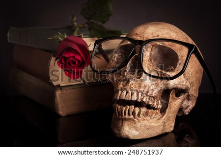 Still life human skull wearing glasses with old book and red rose.