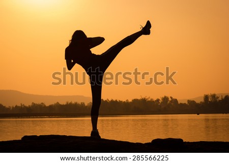 Silhouette of kick boxing girl exercising kick by the river with sunrise background.