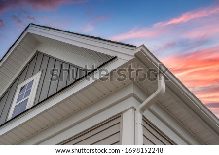 White frame gutter guard system, with gray horizontal and vertical vinyl siding fascia, drip edge, soffit, on a pitched roof attic at a luxury American single family home dramatic sunset sky Foto stock © 