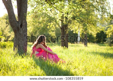 Lovely young woman in a long pink dress sitting on grass under a tree