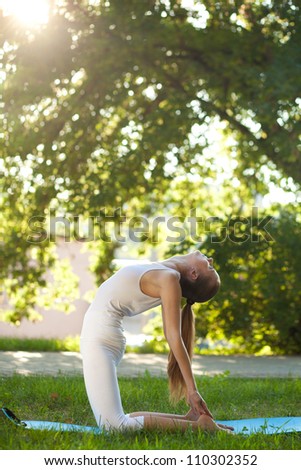 Beautiful young woman practices yoga in nature; standing on her knees, arching back and keeping her feet hands