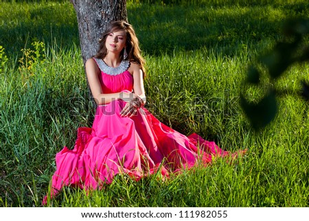Gorgeous young woman in a long pink dress sitting on grass under a tree
