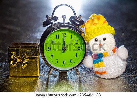 Christmas snowman toy with gold gift box or present and old vintage alarm clock on silver or metal grunge surface. Showing time twelve midnight