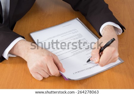 Businessman in dark suit and blue shirt sitting at office desk signing a contract with shallow focus on signature.