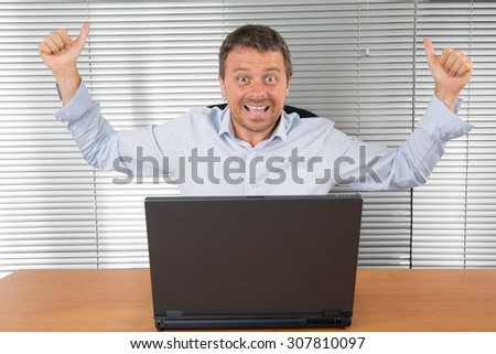 Excited businessman shouting of joy and gesturing with raised clenched fists over successful business deal in front of a computer over dark grey background.