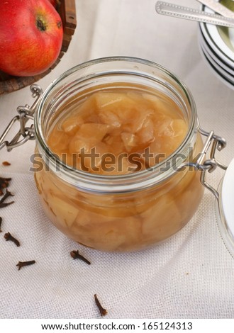 Home made apple compote in a jar (cooked apples in sugar water with spices) on white tablecloth, basket of apples and dried cloves