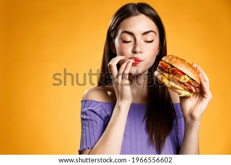 Woman eating cheeseburger with satisfaction. Girl enjoys tasty hamburger takeaway, licking fingers delicious bite of burger, order fastfood delivery while hungry, standing over orange background