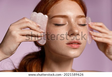 Close up portrait of beautiful redhead girl, ginger female massaging face with jade scraper, close eyes, doing tcm cosmetology procedure, purple background Foto stock © 