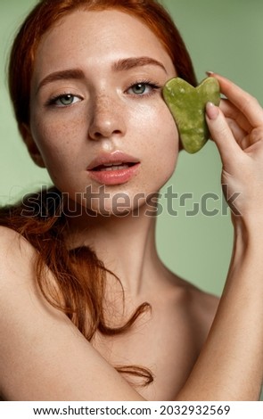 Spa and women beauty. Vertical portrait of redhead girl massage her face with jade massaging scrapper, guasha cosmetology at home, face-lift procedure, standing against green background Foto stock © 
