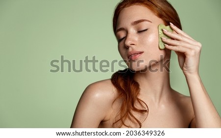 Beautiful redhead woman doing skin care routine after shower, scrapping, massage facial skin with jade gua sha scrapper, body and face beauty concept, green background. Foto stock © 