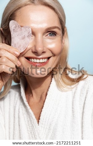 Close up of smiling middle-aged woman with gua sha scrapper massaging tool, using tcm for skin rejuvenation and healthy face, blue background Foto stock © 