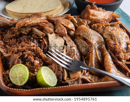 Close up image of a Mexican mud plate with carnitas or pork meat with pork rinds and a fork on top with a sliced lemon and corn tortillas on the side. Mexican carnitas