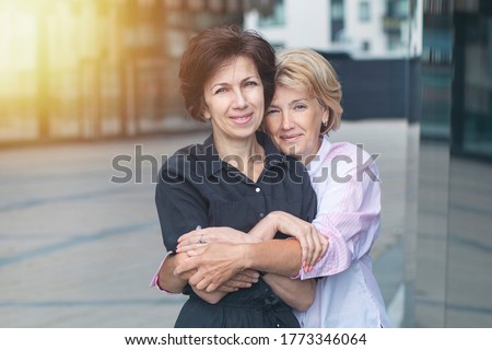 Two happy positive women, female friends standing outdoors office after work. Couple in love hugging, holding each other. Mature senior ladies. LGBT community, lesbian, homosexual, business concept