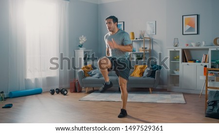 Strong Athletic Fit Man in T-shirt and Shorts is Energetically Jogging in Place at Home in His Spacious and Bright Living Room with Minimalistic Interior. Сток-фото © 