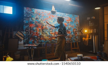 Talented Female Artist Working on a Modern Abstract Oil Painting, Gesturing with Broad Strokes Using Paint Brush. Dark Creative Studio Large Picture Stands on Easel Illuminated, Tools Everywhere