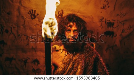 Portrait of Primeval Caveman Wearing Animal Skin Standing in His Cave At Night, Holding Torch with Fire. Primitive Neanderthal Hunter / Homo Sapiens At Night Alone. In the Background Cave Art Drawings Foto stock © 