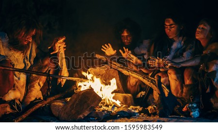 Neanderthal or Homo Sapiens Family Cooking Animal Meat over Bonfire and then Eating it. Tribe of Prehistoric Hunter-Gatherers Wearing Animal Skins Eating in a Dark Scary Cave at Night