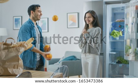 Beautiful Young Couple Have Fun in the Kitchen. Man is Juggling with Oranges. Girl is Clapping, Cheering for Him and They Laugh. Room Has a Modern Tech Fridge with Fresh Groceries. 商業照片 © 