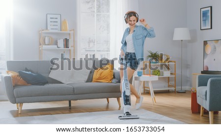 Shot of a Young Beautiful Woman in Jeans Shirt and Shorts Dancing and Vacuum Cleaning a Carpet in a Cozy Room at Home. She Uses a Modern Cordless Vacuum. She's Happy.