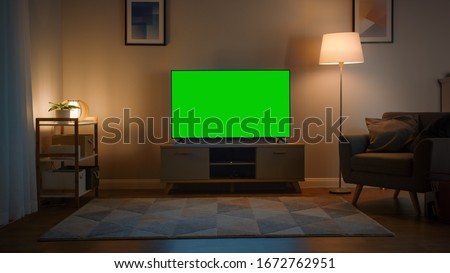 Shot of a TV with Horizontal Green Screen Mock Up. Cozy Evening Living Room with a Chair and Lamps Turned On at Home.