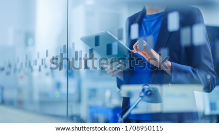 Modern Factory Office: Young and Confident Female Industrial Engineer Standing and Holding Digital Tablet, Using Gestures to Work Efficiently. Focus on Hands and Tablet. Stockfoto © 