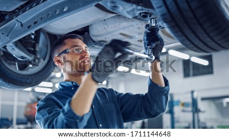 Portrait Shot of a Handsome Mechanic Working on a Vehicle in a Car Service. Professional Repairman is Wearing Gloves and Using a Ratchet Underneath the Car. Modern Clean Workshop. Foto stock © 