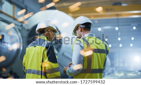 Two Heavy Industry Engineers Stand in Pipe Manufacturing Factory, Use Digital Tablet Computer, Have Discussion. Construction of Oil, Gas and Fuels Transport Pipeline. Back View Sparks Flying 商業照片 © 