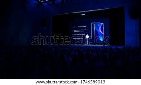 Live Event with Brand New Products Reveal: Keynote Speaker Presents Smartphone Device to Audience. Movie Theater Screen Shows Mock-up Touch Screen Phone with High-Tech Features and Top Highlights