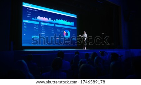 Business Forum Economics Conference Stage: Visionary Female Chief Analyst Delivers Speech and Shows Infographics, Statistics on Movie Theater Screen. Presentation with Speaker in Full Auditorium Hall