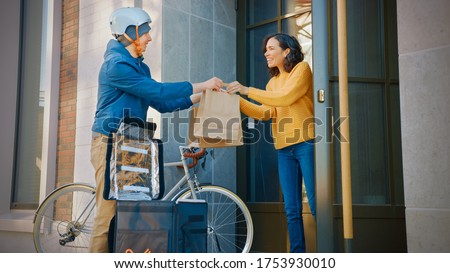 Happy Food Delivery Man Wearing Thermal Backpack on a Bike Delivers Restaurant Order to a Beautiful Female Customer. Courier Delivers Takeaway Lunch to Gorgeous Girl in Urban Building.